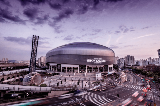 The Gocheok Sky Dome in Seoul, South Korea. A dome-shaped stadium with a large flagpole in front of a multi-lane road, with the city in the background.