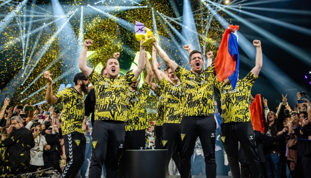 ZywOo stands at the front of the stage to lift the BLAST Paris trophy with Team Vitality. Confetti is falling in the background, and his teammate holds a French flag.