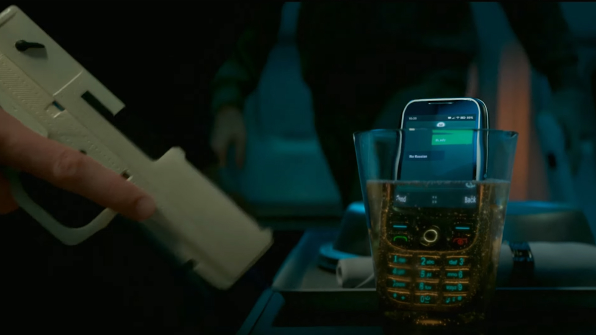 An old mobile phone sits in a small glass with liquid on a plane, as two terrorists prepare weapons in Call of Duty: Modern Warfare 2.