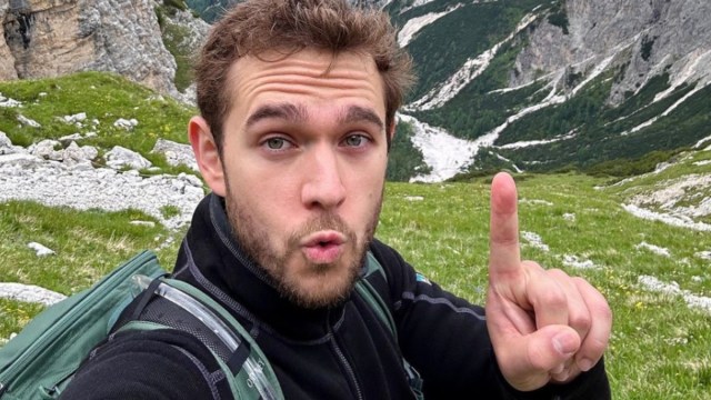 Zedd pointing his finger up in a selfie in front of mountains