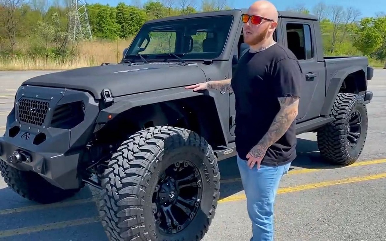 There is a picture of TimTheTatMan standing in front of his Jeep. The Jeep is black and has trees in the background behind it. 