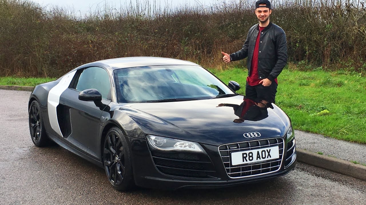 Theere is a picture of Ali-A standing next to his car. The car is black. They are both in the side of the road. 