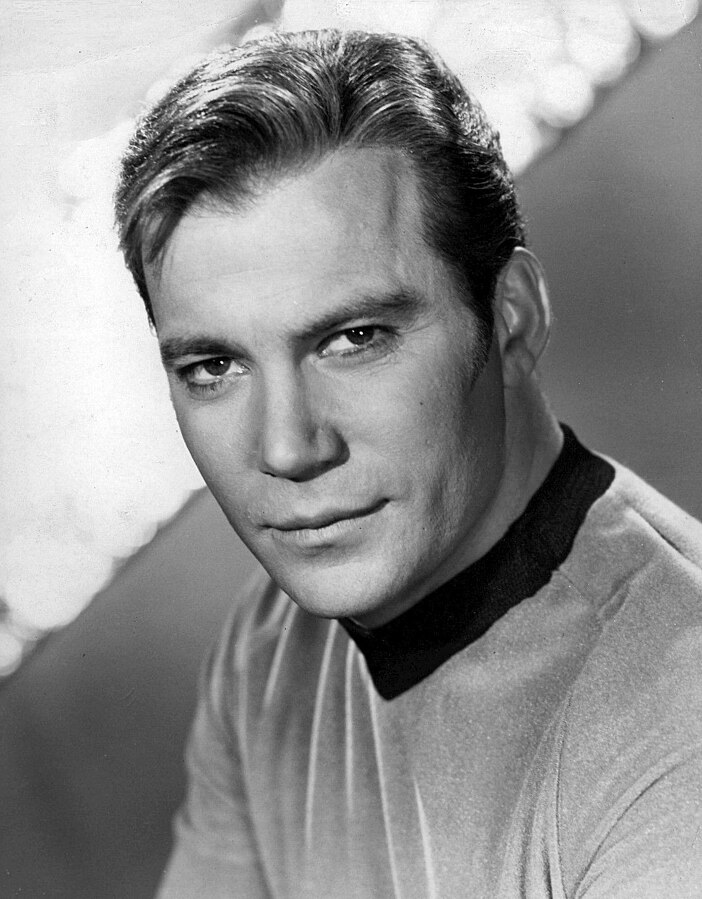 A black and white image of William Shatner in costume as Captain James T Kirk from Star Trek. 