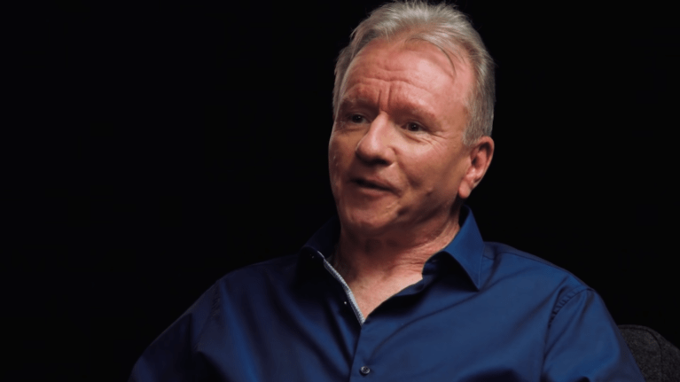 Jim Ryan is a white caucasian man with gray short hair and no faical hair. The image is a close up on Jim during an interview and he is wearing a blue buttun up shirt.