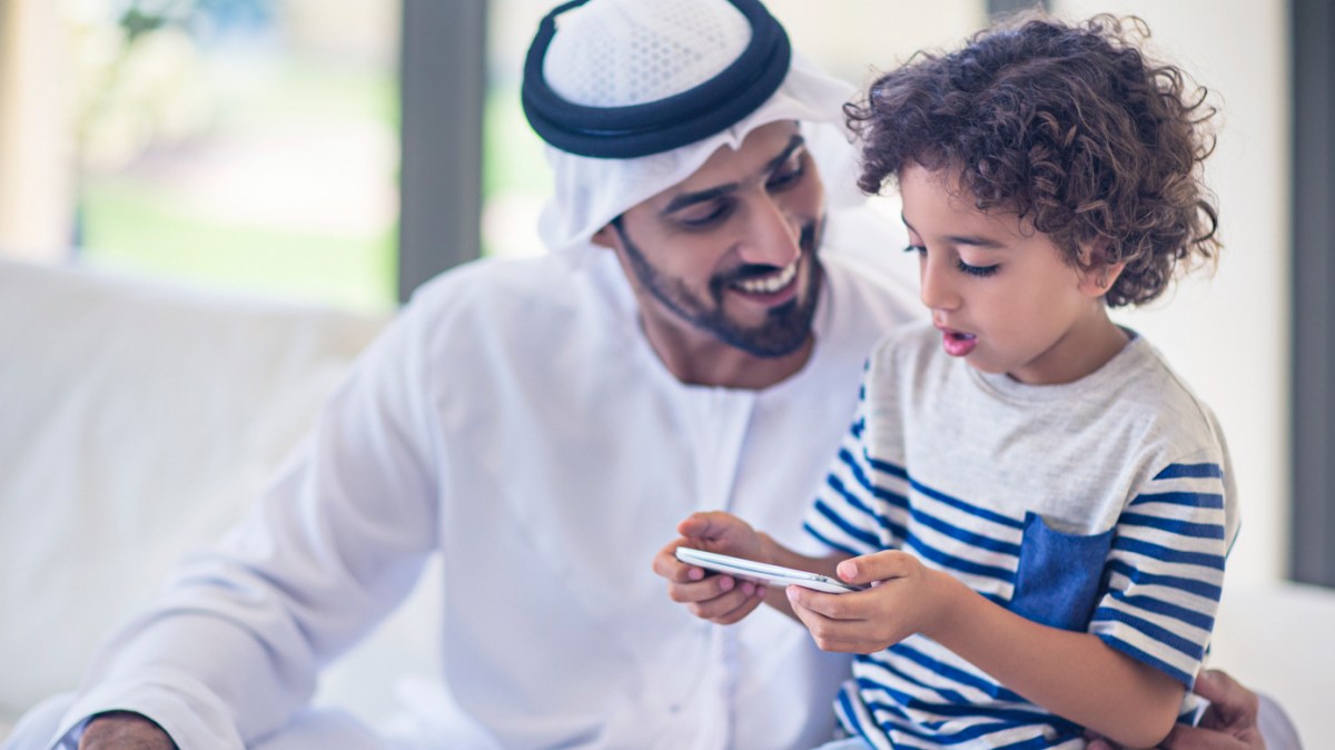 Middle Eastern father sharing time with his son at home, kid is playing with the mobile phone while his father is looking at him,the father is wearing the traditional Dishdasha.