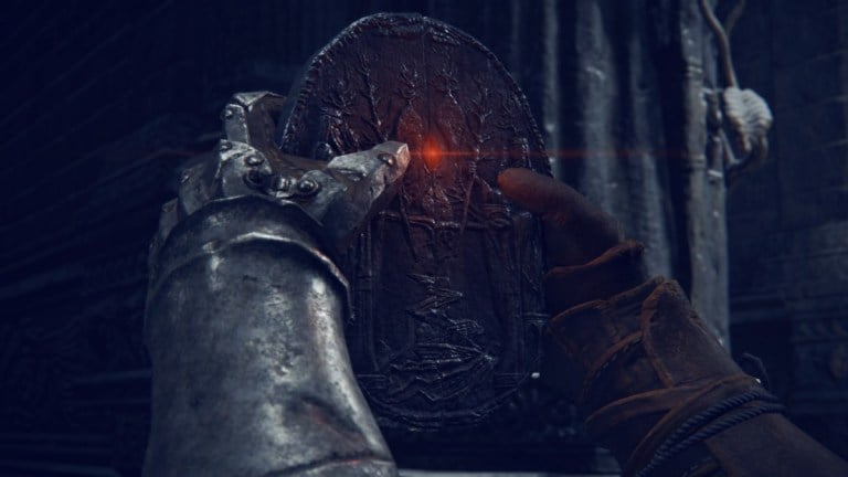 A pair of armored gloves hold up a stone medallion with a red gemstone sample in Elden Ring.