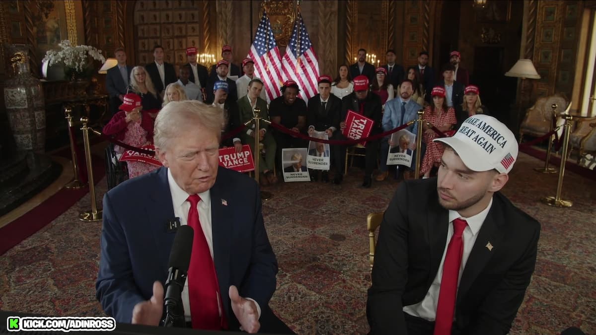 Trump on Adin Ross stream, sitting down in a suit with a red tie and bystanders sitting in the background.