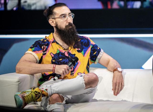 CS2 talent Jacky sitting on a white couch looking at someone off-screen while holding a microphone close to his yellow panther shirt.