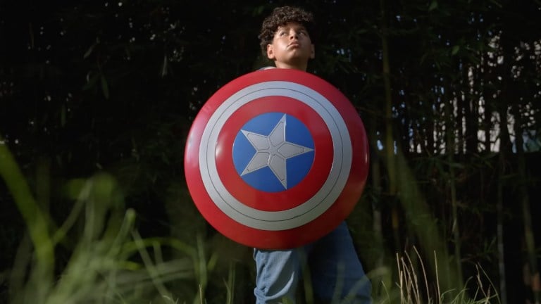 A boy holds Captain America's red, white, and blue shield while standing in long grass.