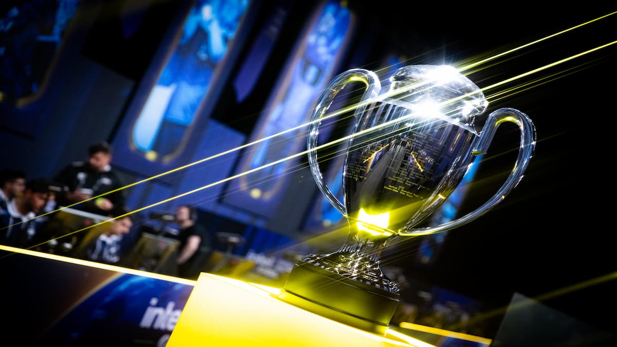 A silver trophy sitting on a yellow pedestal. There are people and screens in the background.